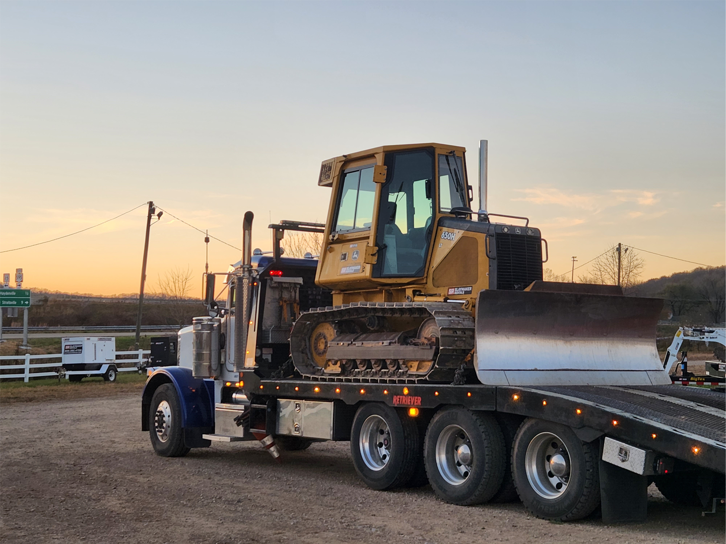 Freight truck carrying bulldozer in Slaymaker Rentals yard at dawn